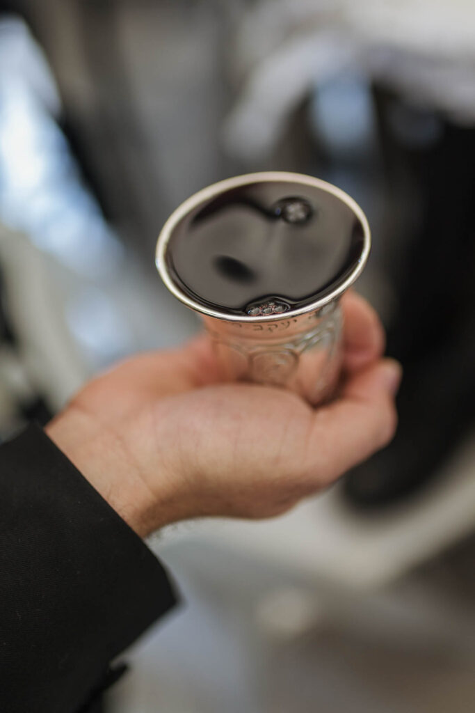 כוס יין כסף מלאה ביין אדום, מוחזקת ביד במהלך טקס ברית מילה, רקע מטושטש